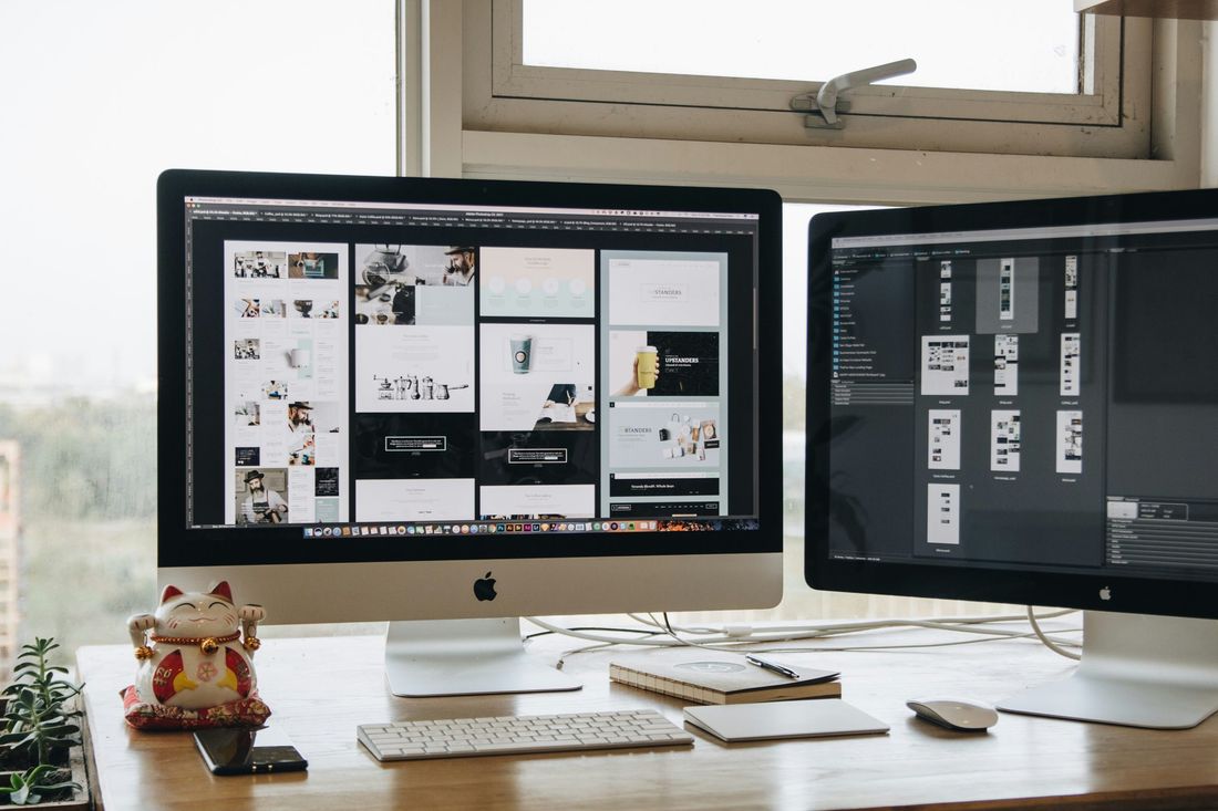Two monitors on a desk showing website page layouts and design files